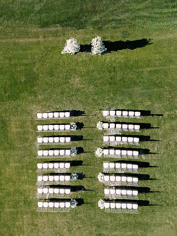 Ceremony seating layout with white chairs lining a center aisle, featuring floral arrangements at aisle ends on a grass lawn