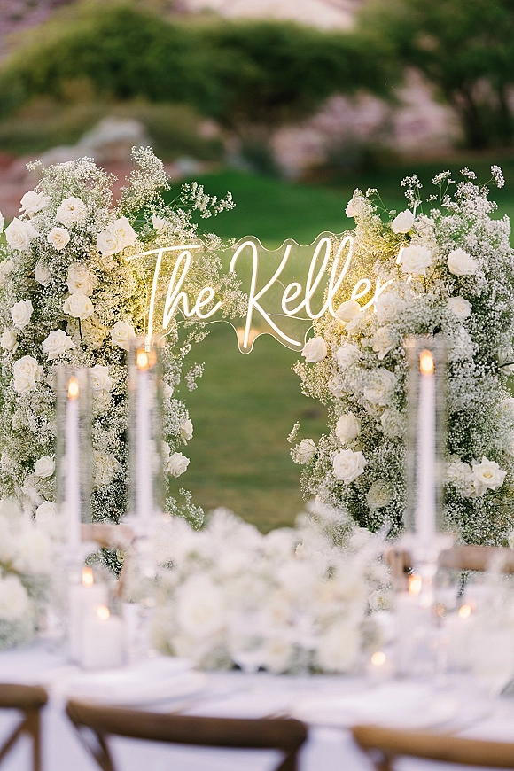 Wedding neon sign glowing above a white rose and baby’s breath floral arch, with taper candles on the table on a garden lawn backdrop