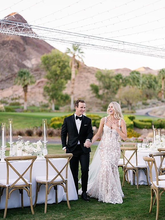 Couple portrait of bride and groom holding hands, walking past outdoor reception tables under string lights with mountains and palm trees behind