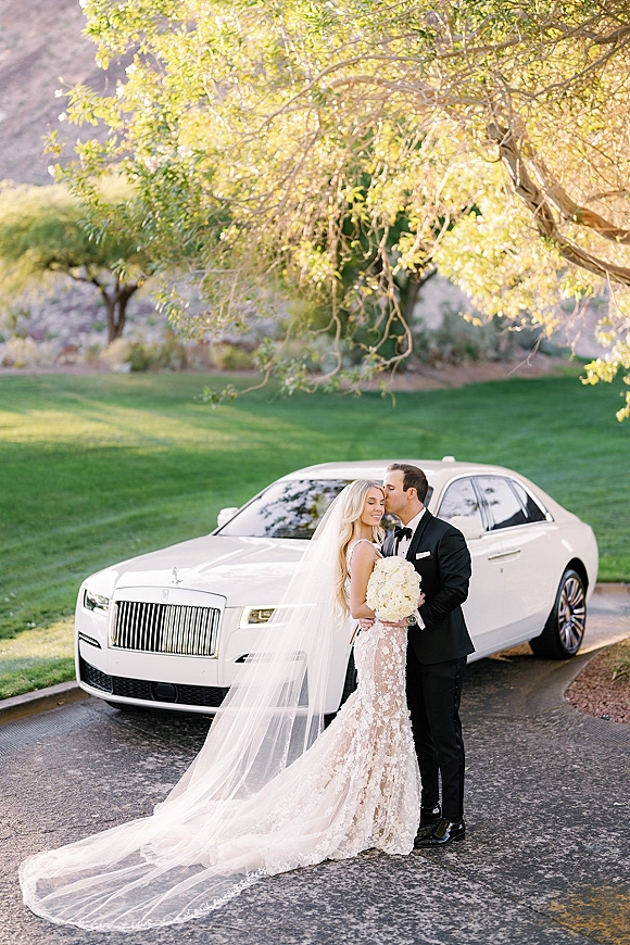 Couple portrait of bride and groom kissing by a white luxury car, her lace dress and long veil trailing, holding a white bouquet on a green lawn