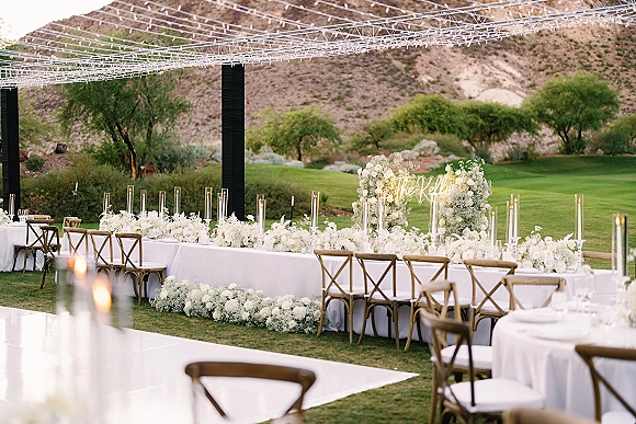 Reception tablescape with long head table decor, white floral centerpieces and tall cylinder candles under an overhead canopy on a lawn with desert mountains beyond