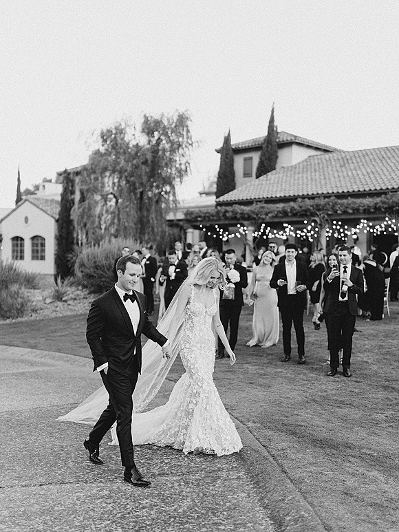 Wedding exit as bride and groom walk away holding hands under string lights at dusk, guests lining the walkway near a villa lawn