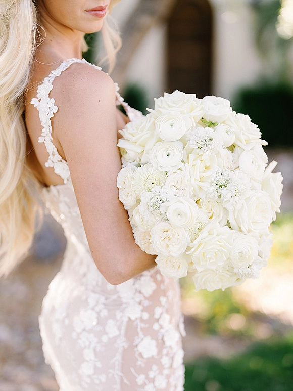 Bridal bouquet of white roses and ranunculus held against a lace wedding dress with floral appliqué straps in a blurred garden setting