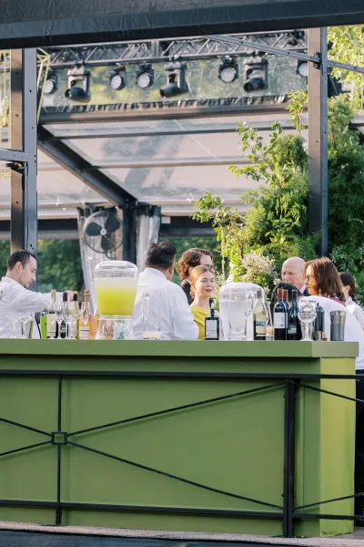 Wedding bar setup with an outdoor wedding bar counter, glass drink dispensers and bottles under a glass pavilion with greenery and guests