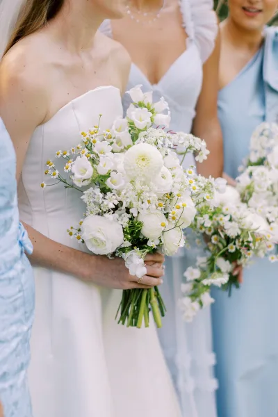 Bridesmaid bouquets of white blooms and greenery, with chamomile daisies, held by bridesmaids in blue dresses against outdoor greenery