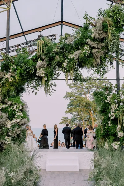 Wedding ceremony decor with a floral arch wedding of white blooms and greenery garland under a glass roof, with ocean view and guests nearby