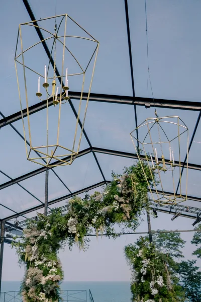 Wedding ceremony decor with hanging chandelier and taper candles above a lush white floral arch under a clear-top tent with trees beyond