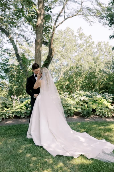 Couple portrait of bride and groom embrace as she touches his face, cathedral veil trailing over grass in dappled garden light