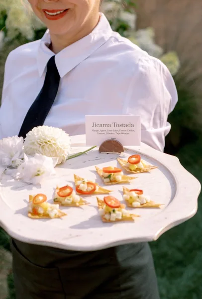 Wedding passed appetizers on a white tray with mini tostadas, jicama and sliced peppers, held by a server in sunlit garden greenery