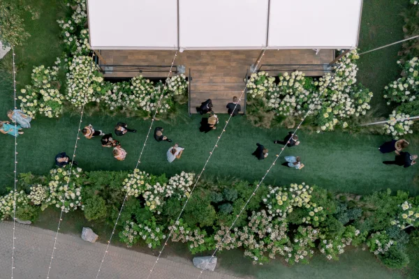 Outdoor wedding reception under a string lights canopy, with white flowers and greenery bordering a lawn walkway beside a tented stage
