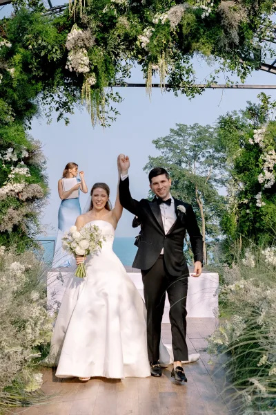 Recessional moment as bride and groom recessional raise hands, bouquet and tuxedo under greenery floral arch by water and trees