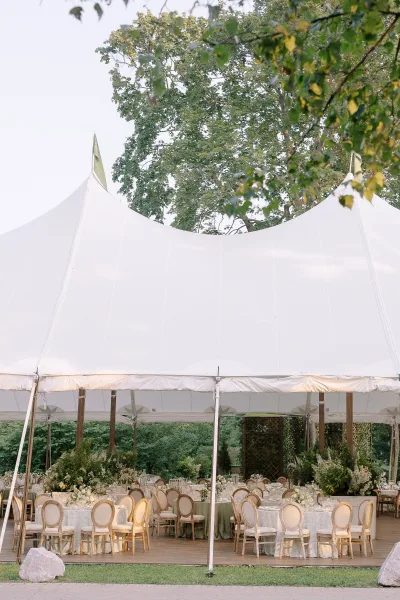 Reception tent decor in a sailcloth wedding tent with round tables, green linens, upholstered chairs, and lush greenery centerpieces on a wood deck