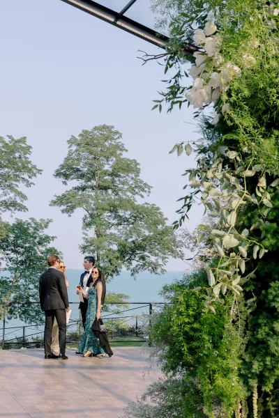 Wedding cocktail hour with wedding guests mingling on an outdoor terrace, holding drinks near a floral arch with lake view beyond railing