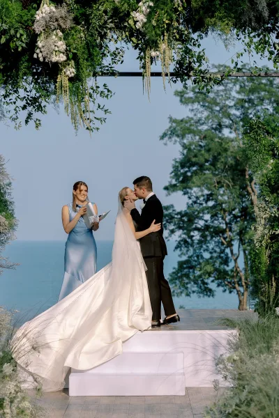 Ceremony kiss under a floral arch, bride’s veil over face and long train gown as groom in tux kisses with ocean view behind