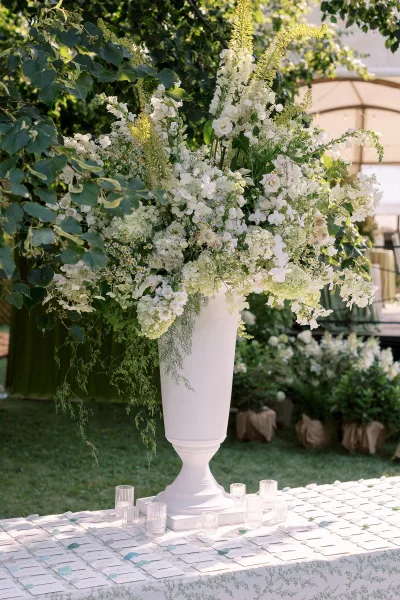 Wedding floral centerpiece in a white urn centerpiece with lush white blooms and greenery, ribbed glass votives and escort cards under a tented lawn setting