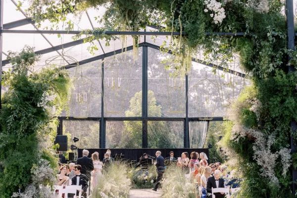 Ceremony setup in a greenhouse wedding ceremony with hanging greenery, candle chandeliers, white chairs, and a stage framed by glass walls