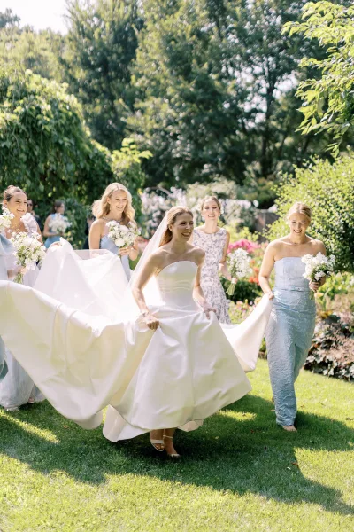 Bride with bridesmaids in a garden, laughing as they lift her long train and carry white bouquets, veil trailing over the lawn