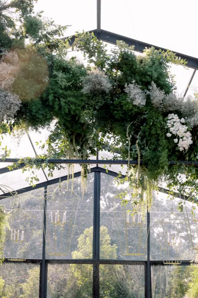 Hanging floral installation with greenery garland and white flowers drapes from geometric frames with candelabra candles beneath a glass greenhouse ceiling