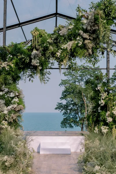 Wedding ceremony arch with greenery wedding arch and white hanging florals inside a glass greenhouse, with ocean view beyond and bench seating