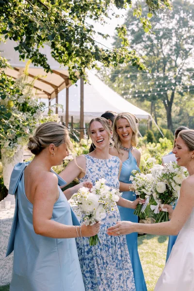 Bridesmaid group photo of bridesmaids holding bouquets in blue dresses, smiling on a sunny lawn by a floral wedding tent