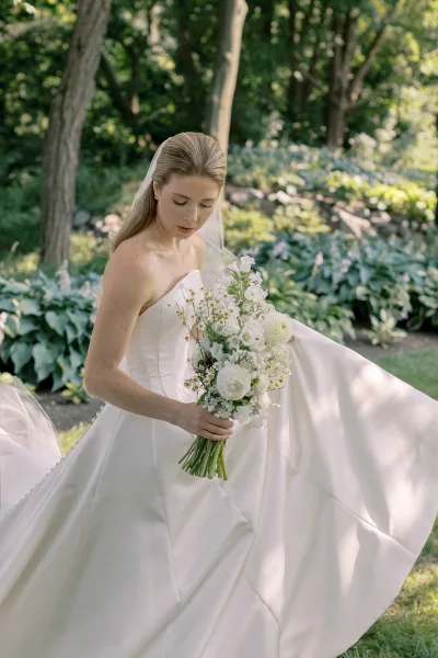 Bridal portrait of a bride looking down in a strapless satin wedding dress with long veil, holding a white bouquet in a garden setting