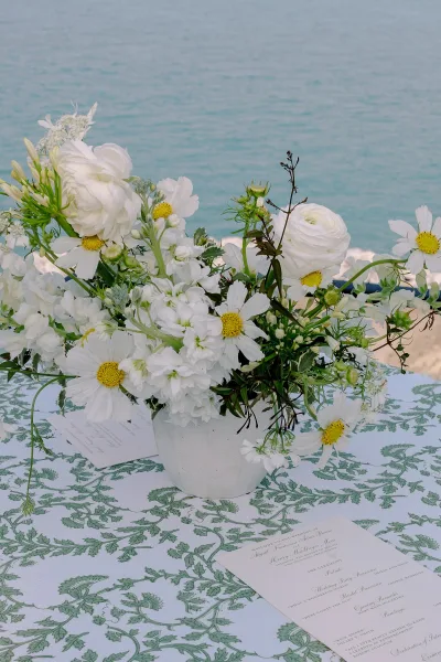 Wedding centerpiece with a white floral centerpiece in a ceramic vase and greenery on a patterned tablecloth, set beside the ocean shoreline