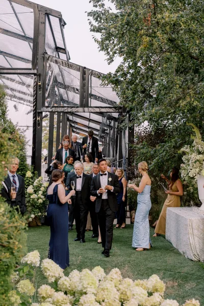 Wedding cocktail hour with wedding guests mingling in tuxedos and dresses, holding champagne flutes near hydrangeas by a glass greenhouse