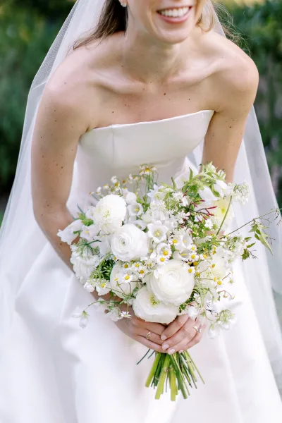 Bridal bouquet of white flowers and greenery in the bride’s hands, bridal veil and strapless dress visible against outdoor greenery