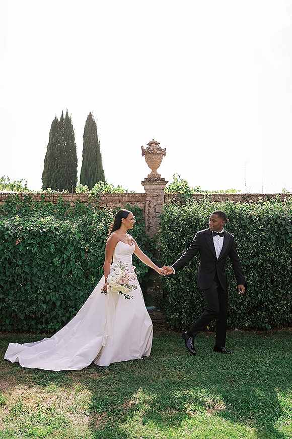 Couple portrait of bride and groom holding hands, bride in strapless gown with train and bouquet by an ivy-covered stone garden wall.