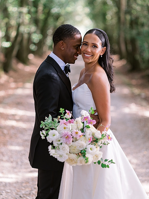 Couple portrait of bride and groom embrace as he kisses her cheek; she smiles holding a white and pink bouquet on a tree-lined path