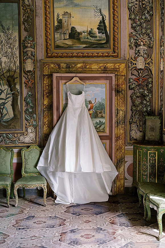 Wedding dress, satin wedding dress hanging on a hanger against ornate painted wall panels with gilded frame, antique green chairs, mosaic floor