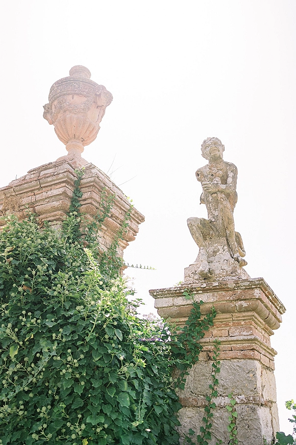 Garden statues and stone garden statue on pedestals beside an antique urn, framed by brick pillars with ivy vines against greenery