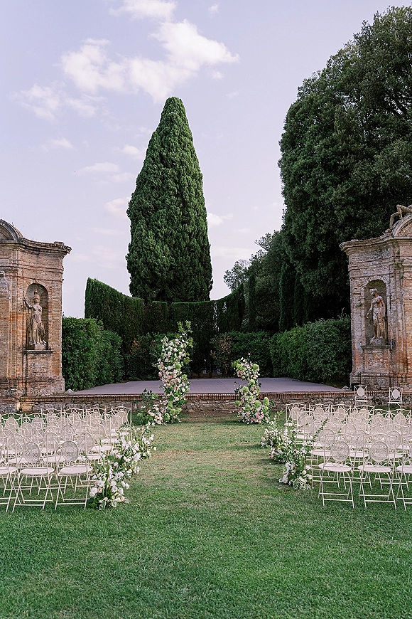 Outdoor ceremony setup with garden wedding ceremony floral pillars and aisle florals, white wrought iron chairs on a manicured lawn by hedges and stone statues