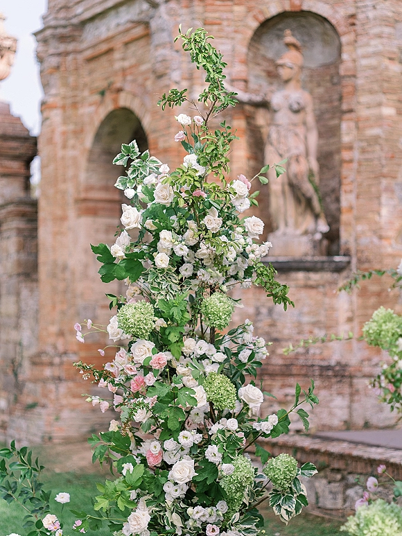 Wedding floral installation with white roses and blush flowers cascading over a pillar, set against a brick arch wall and garden lawn