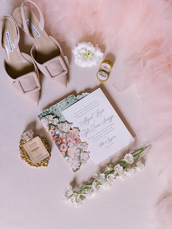 Invitation suite wedding invitation flat lay with floral envelope liner, rings, blush heels, perfume, and white flowers on neutral fabric