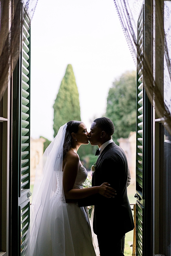 Wedding kiss portrait of bride and groom kissing in window light, her veil and bouquet silhouetted by green shutters and lace curtains