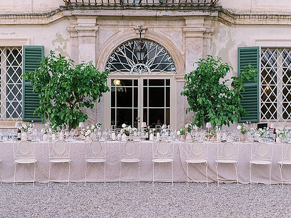 Reception tablescape with long banquet table wedding details—neutral linens, white metal chairs, bud vases, greenery, lamps and wine in a stone villa courtyard