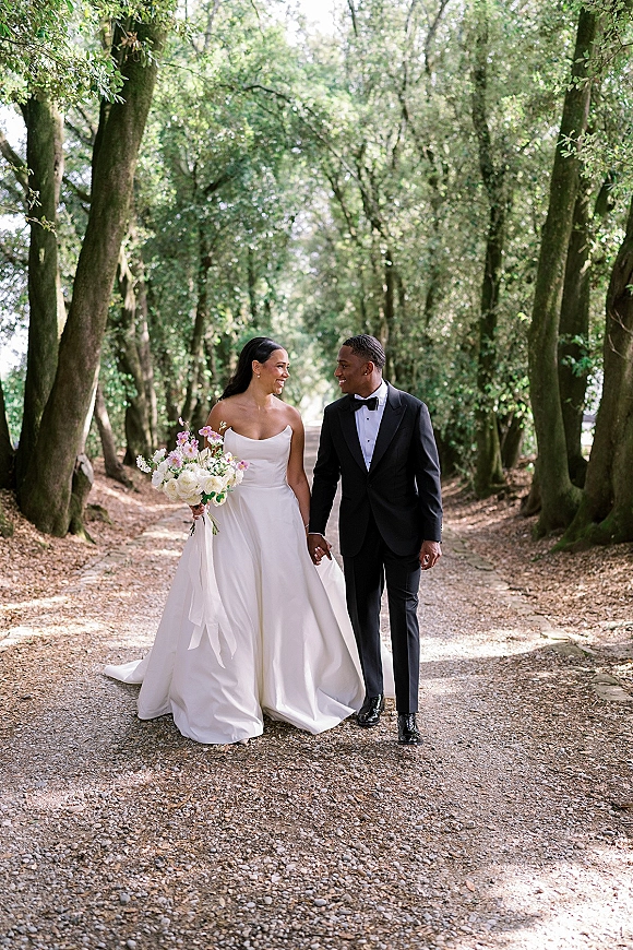 Couple portrait of bride and groom walking hand in hand, bouquet and strapless gown with train on a tree-lined gravel path