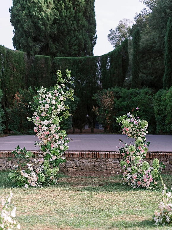 Wedding ceremony arch with asymmetrical floral pillars of roses, hydrangeas, and greenery on a garden lawn against stone wall and hedges