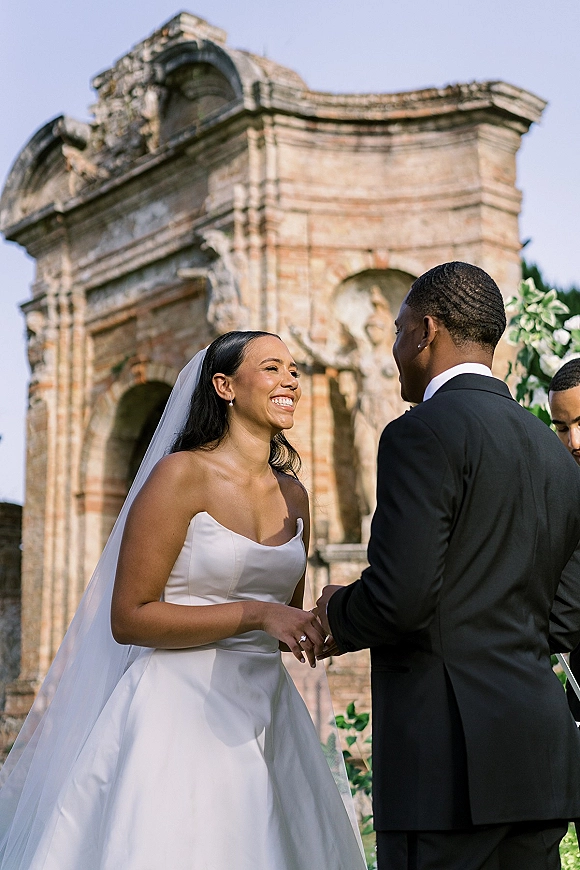 Wedding vows as bride in a strapless dress and veil holds the groom’s hand under a stone archway with white flowers and blue sky