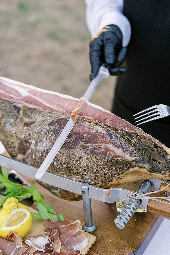 Prosciutto carving station with cured ham leg on a stand, black-gloved hands slicing thin prosciutto on a wooden board outdoors