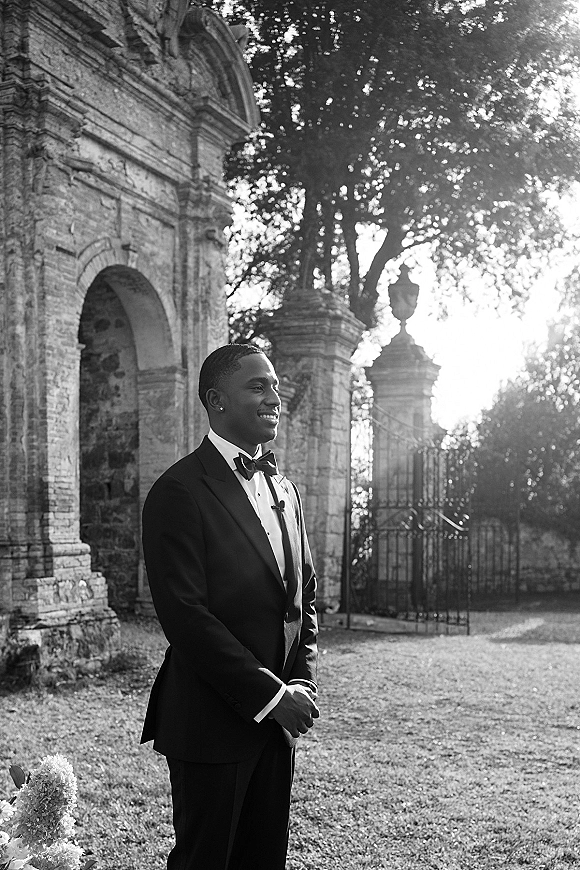 Groom portrait of a black tie groom in a tuxedo and bow tie with boutonniere, standing by a sunlit stone arch and iron gate