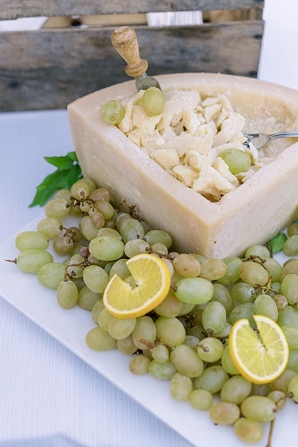 Cheese wheel display with parmesan shavings, green grapes, and lemon slices on a platter, set on a white tablecloth against wood backdrop
