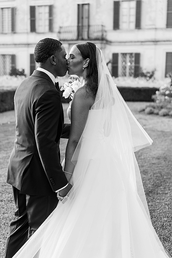 Wedding kiss portrait in black and white of bride and groom kissing in side profile, her long veil flowing before an estate facade and garden lawn