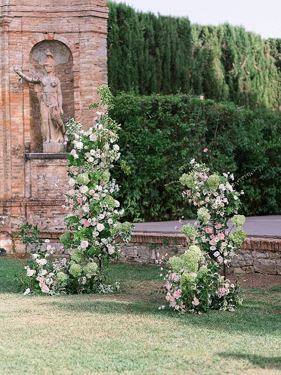 Ceremony floral arrangements of roses and hydrangea with greenery on tall stands, set before a stone wall with statue and hedges