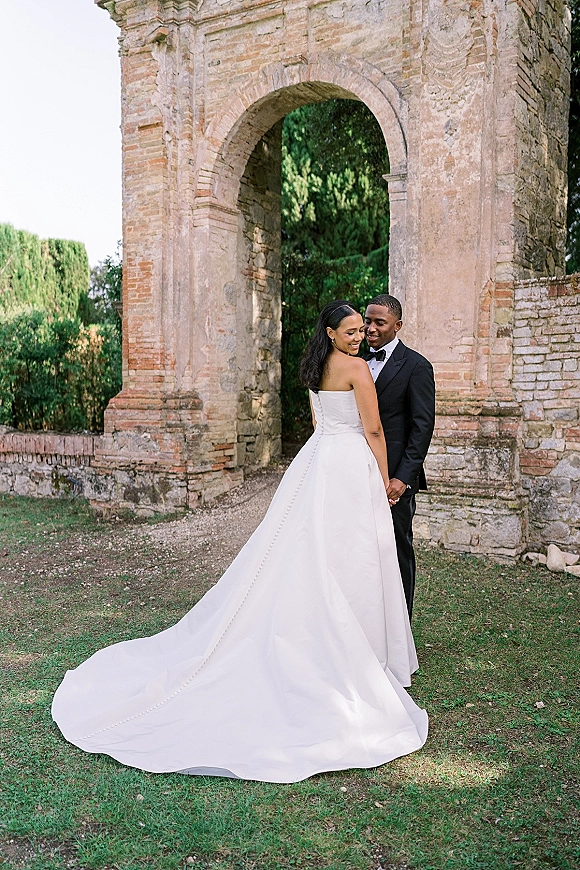 Couple portrait of bride in a strapless gown with long train and groom in tuxedo holding hands beneath a stone archway and brick wall