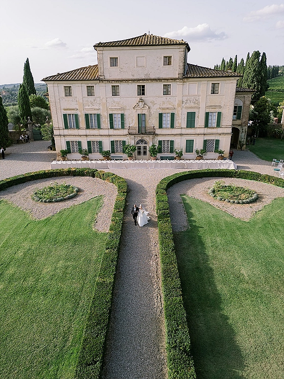 Wedding venue exterior with a long banquet table in white linens set in a gravel courtyard before a historic villa and cypress-lined gardens