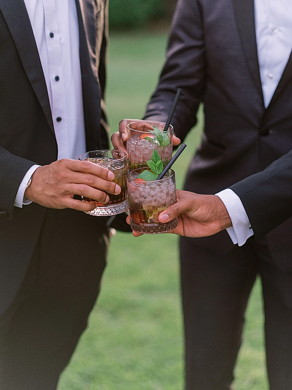 Wedding cocktail toast as groomsmen in tuxedos clink three garnished drinks with ice and straws on an outdoor lawn during cocktail hour