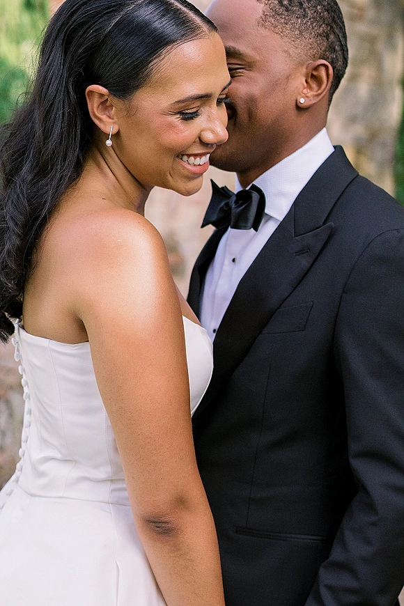 Wedding couple portrait of groom whispering as the smiling bride in a strapless dress hugs him, outdoors by greenery and a stone wall
