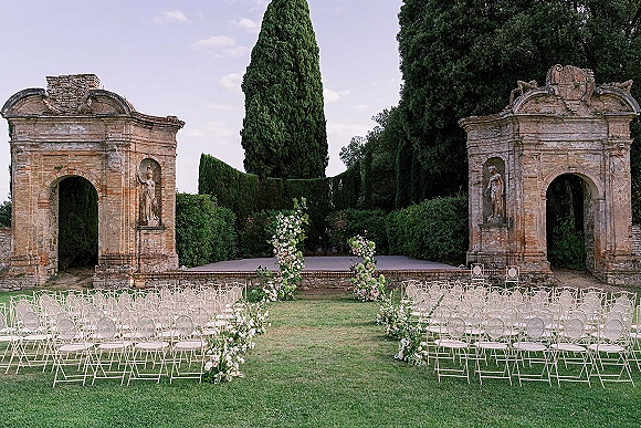 Ceremony setup for an outdoor wedding ceremony with white metal chairs lining a floral aisle toward stone arches on a manicured lawn